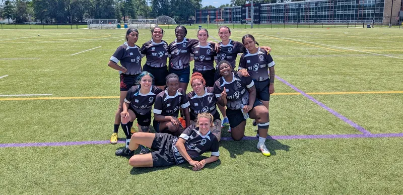 The Women's+ 7s squad posed together on a sunny turf pitch in black Pig N' Whistle jerseys, one player lying in front, a school building behind them