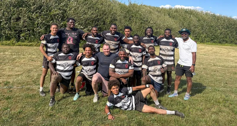 The Men's 7s squad posed together on a grass pitch in black and white hooped jerseys, a hedgerow behind them