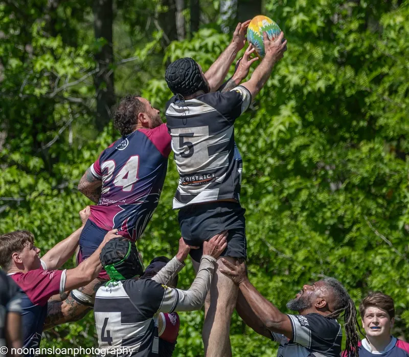 A Lions jumper in a scrum cap takes lineout ball at full stretch, lifted by two teammates and contested by an opposition jumper in maroon