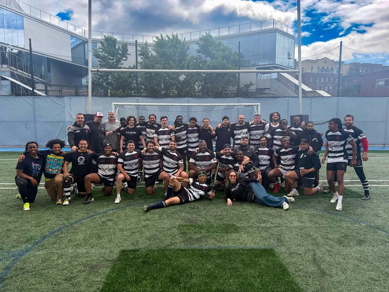 The Men's 15s squad and supporters gathered in front of the posts on a turf pitch, city buildings behind
